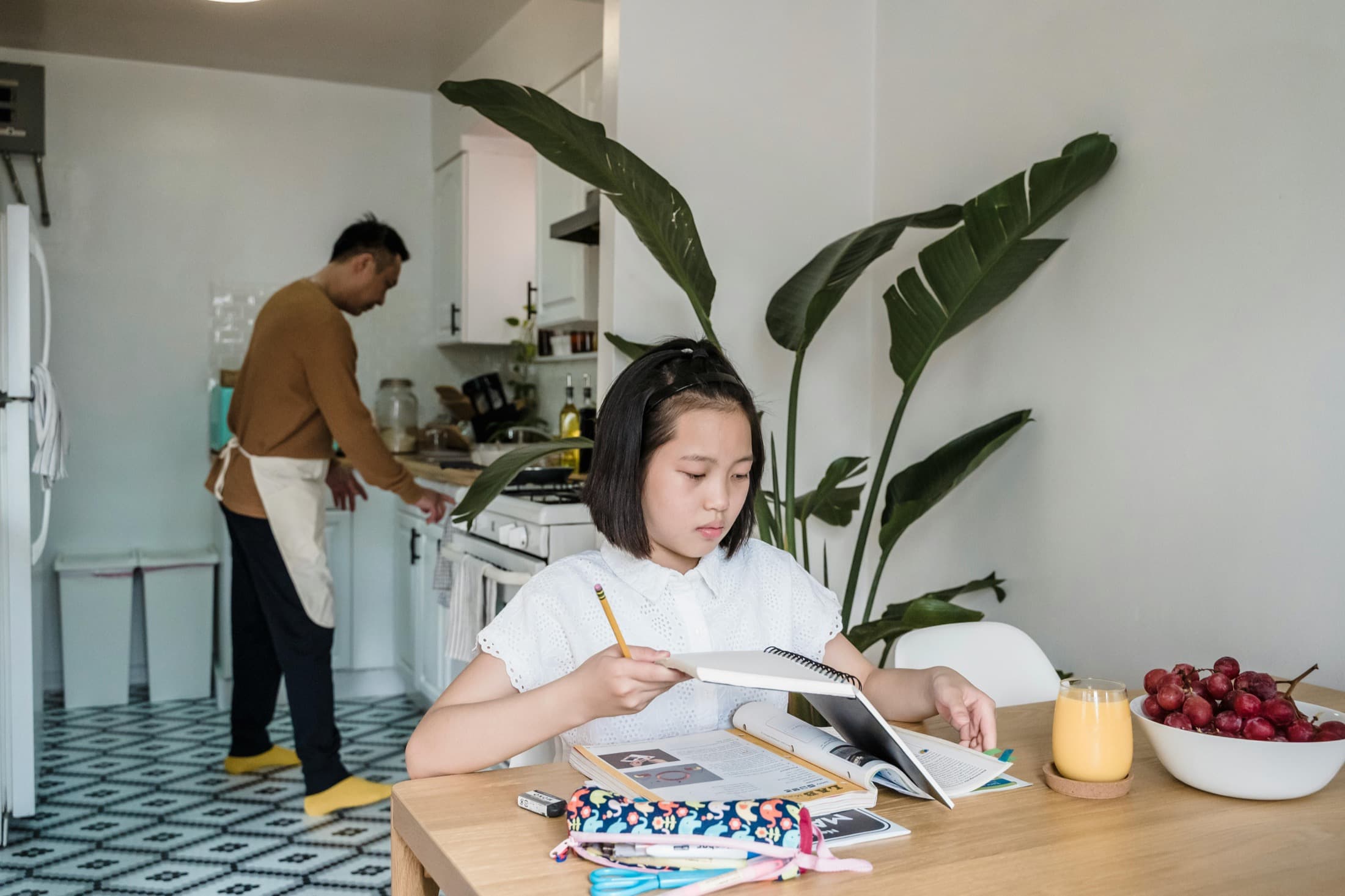 A child doing homework at a dining table while a parent works in the kitchen