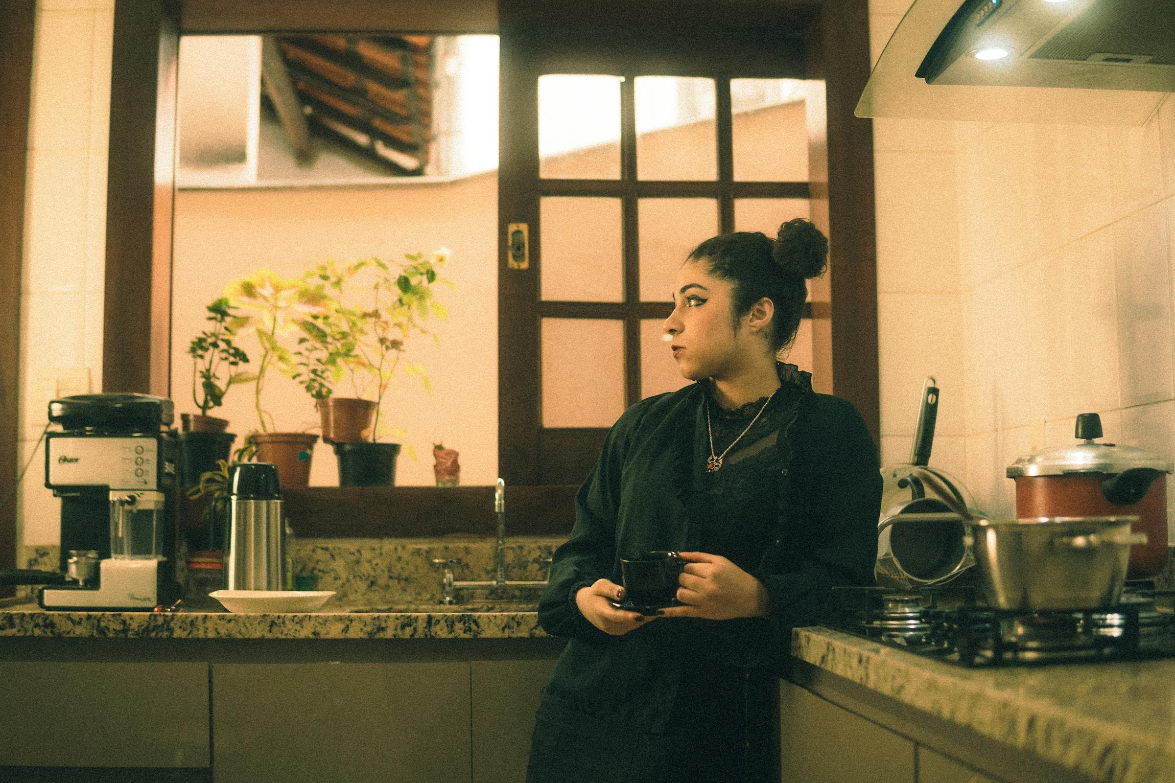 A woman alone in her kitchen at morning, holding a coffee cup, looking out the window