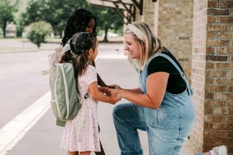 A parent crouched at a school gate with a child in a backpack