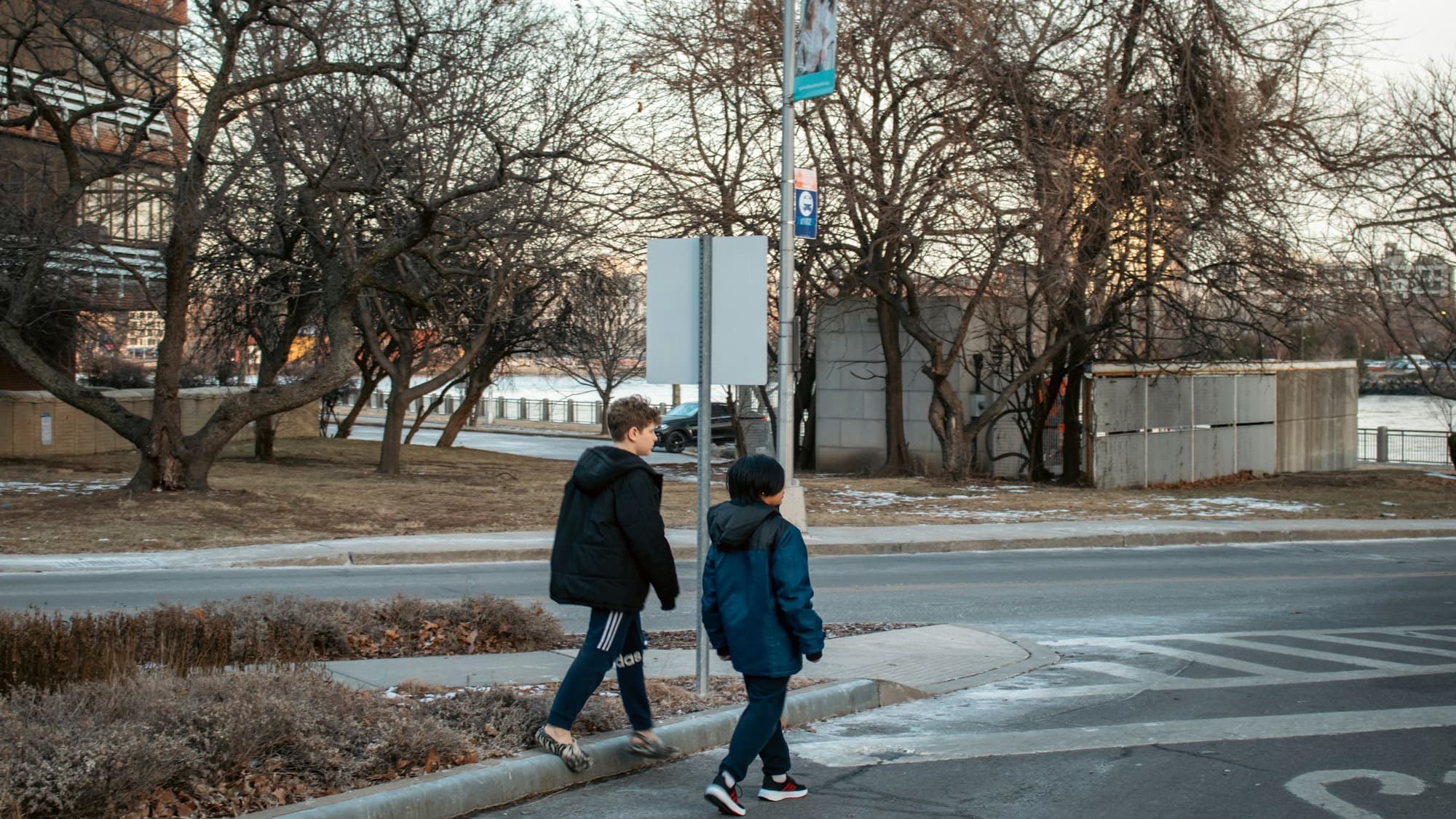 Two kids in winter coats walking together along a city street toward a crosswalk
