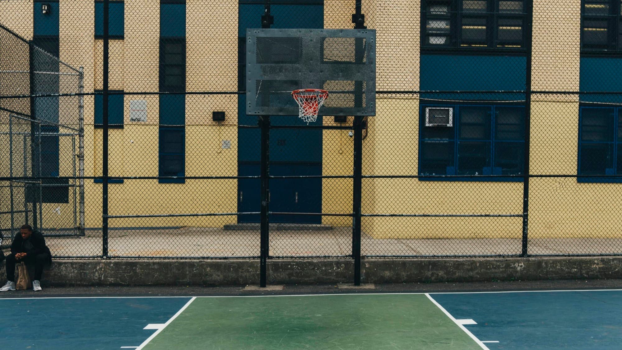 An empty schoolyard basketball court behind a chain-link fence, with one person sitting alone at the edge